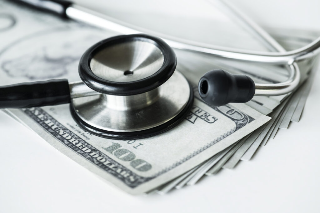 Close-up of a stethoscope resting on a stack of hundred-dollar bills, symbolizing healthcare costs.