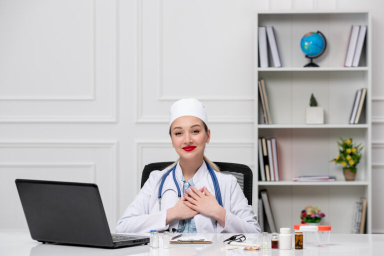 A smiling healthcare professional sitting at a desk with medical supplies and a laptop in a clinical office setting.