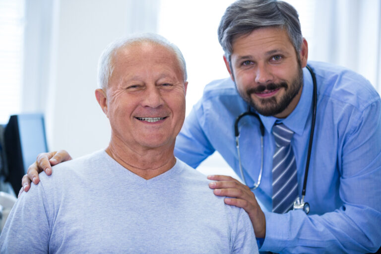 A senior man smiling beside a friendly healthcare professional in a medical office.